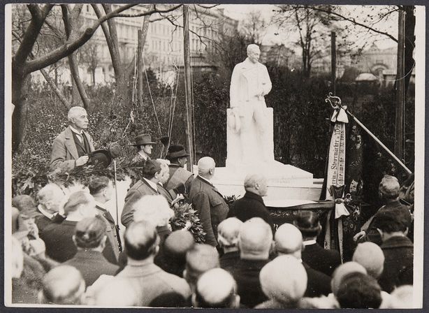 Black and white photograph of a ceremony in a park. A group of people in period dress stand around a newly unveiled statue of a man. Some people carry bouquets of flowers, and in the foreground an elderly man holds a hat in his hand. A ribbon with lettering is attached next to the statue.